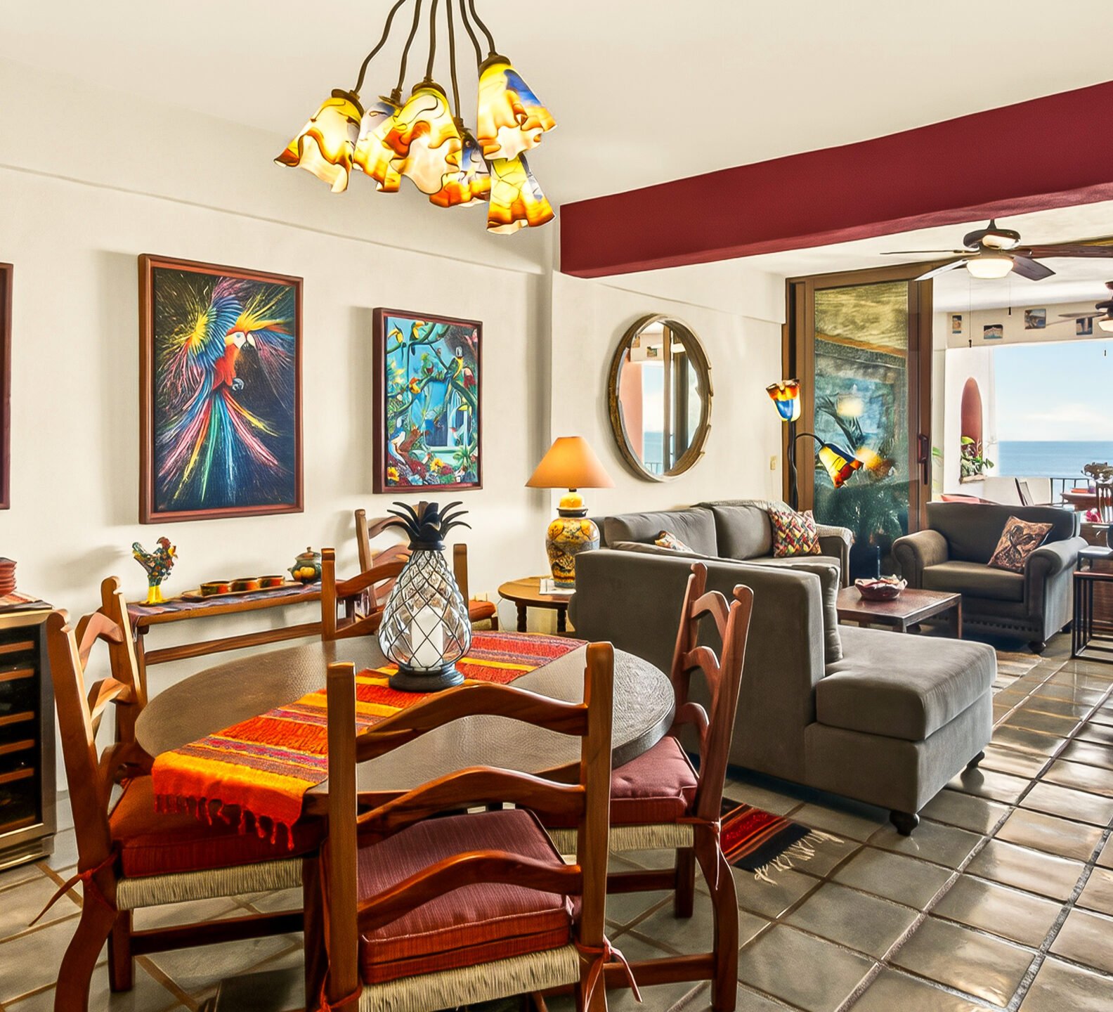 A brightly lit, open-concept dining and living area with rustic tile flooring that opens up to an outdoor patio with an ocean view.